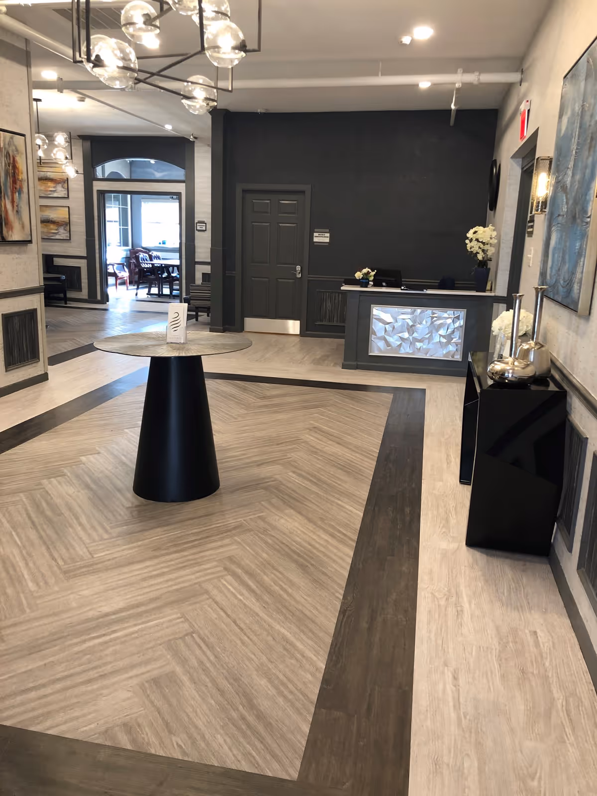 Modern lobby reception area with a central round table, front desk, seating visible in the background, and decorative lighting.