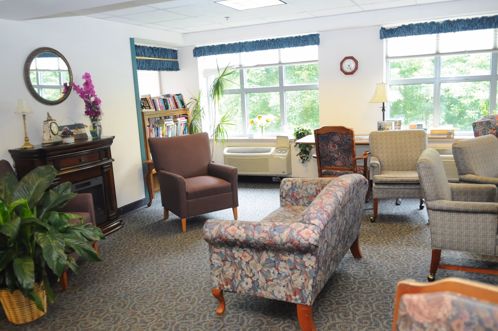 A cozy living room area with various upholstered chairs and a floral-patterned sofa arranged on a carpeted floor. There is a wooden cabinet with a clock and lamp on top, a round mirror on the wall, a bookshelf filled with books, and large windows letting in natural light. A potted plant is visible in the corner.