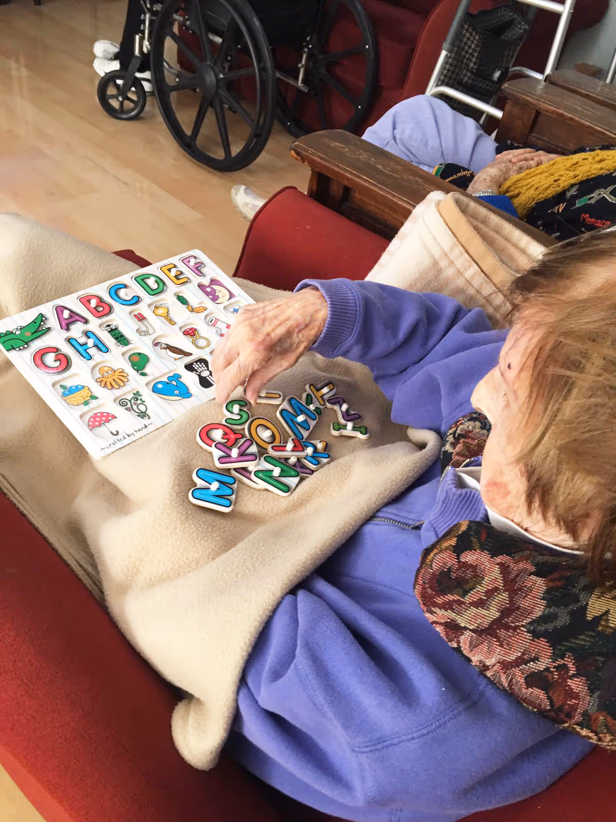 An elderly person sitting in a red armchair covered with a beige blanket, playing with colorful alphabet letters and a matching alphabet board. A wheelchair is visible in the background on a wooden floor.