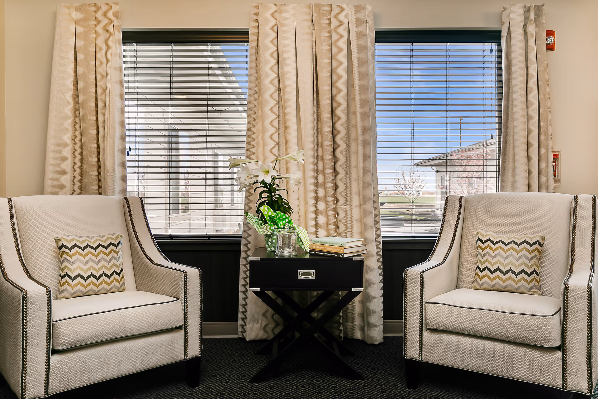 Two beige upholstered armchairs with patterned throw pillows are positioned on either side of a small black side table. The table holds a potted white lily plant wrapped in green polka dot paper, a glass candle holder, and a stack of books. Behind the chairs are two large windows with beige patterned curtains and horizontal blinds, showing an outdoor view of a building and trees.