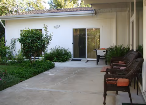 Concrete patio outside a white single-story building with wicker chairs, potted plants, and a sliding glass door.