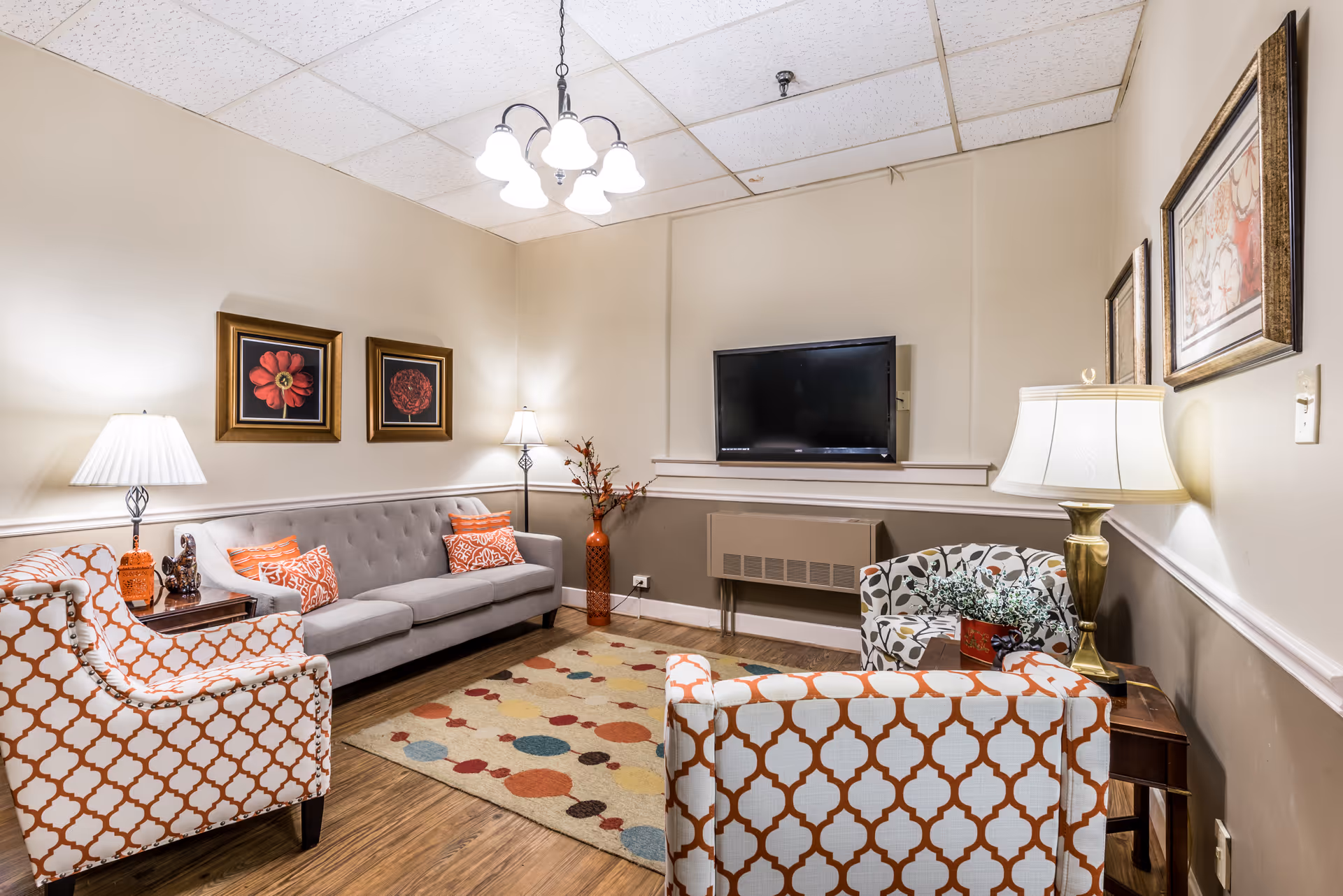 A cozy living room with a gray sofa adorned with orange patterned pillows, two armchairs with orange and white geometric patterns, a colorful rug with circular designs, a wall-mounted flat-screen TV, and several lamps providing warm lighting. The walls are decorated with framed floral artwork.