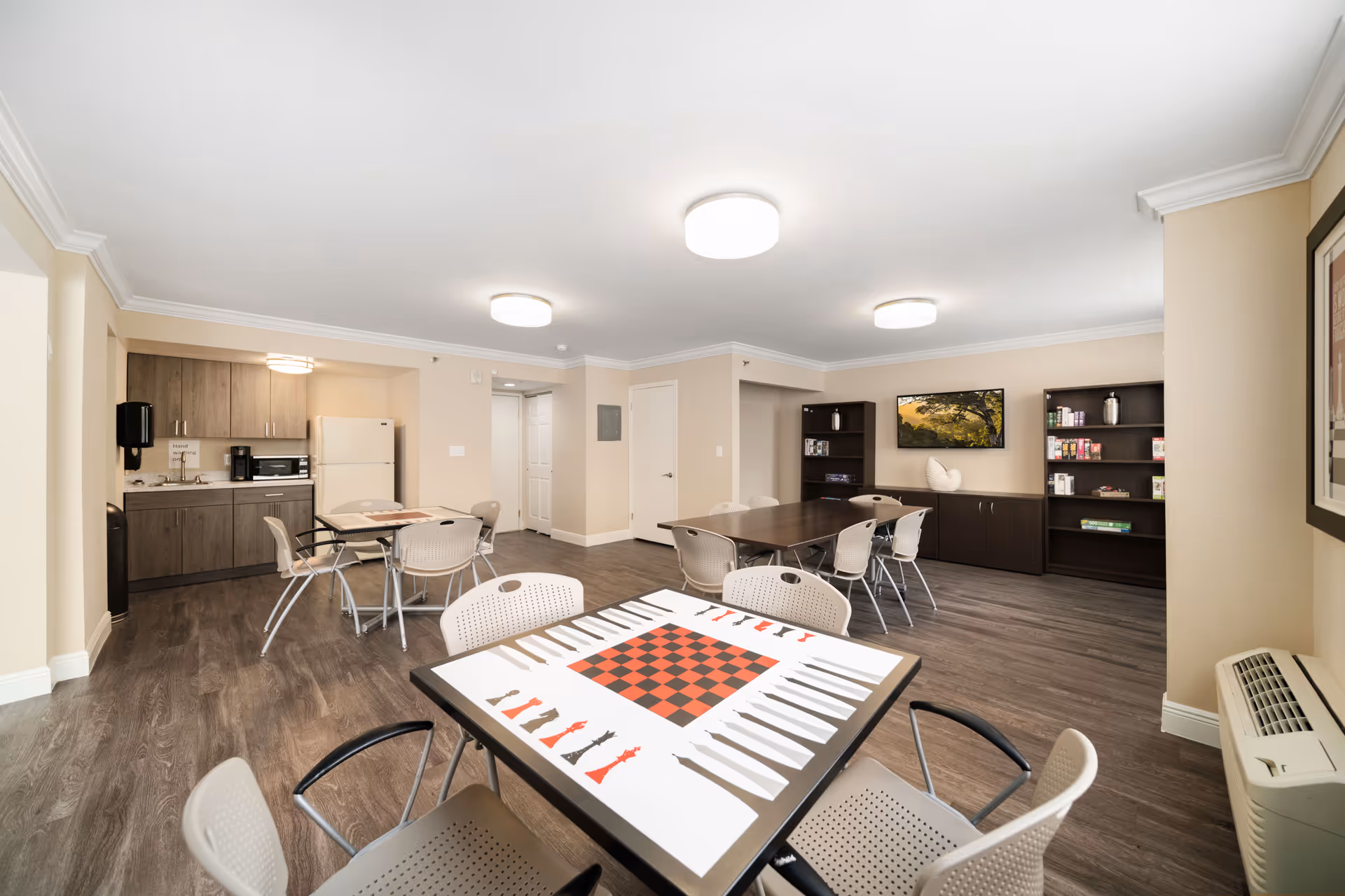 A spacious common room with light beige walls and wood flooring. The room features multiple tables and chairs, including a table with a built-in chess and checkers board in the foreground. There are two dark wood bookshelves and a cabinet against the far wall, with a flat-screen TV mounted above the cabinet displaying a nature scene. To the left, there is a small kitchenette area with a refrigerator, microwave, coffee maker, and sink. The room is well-lit with ceiling lights and has a clean, modern appearance.