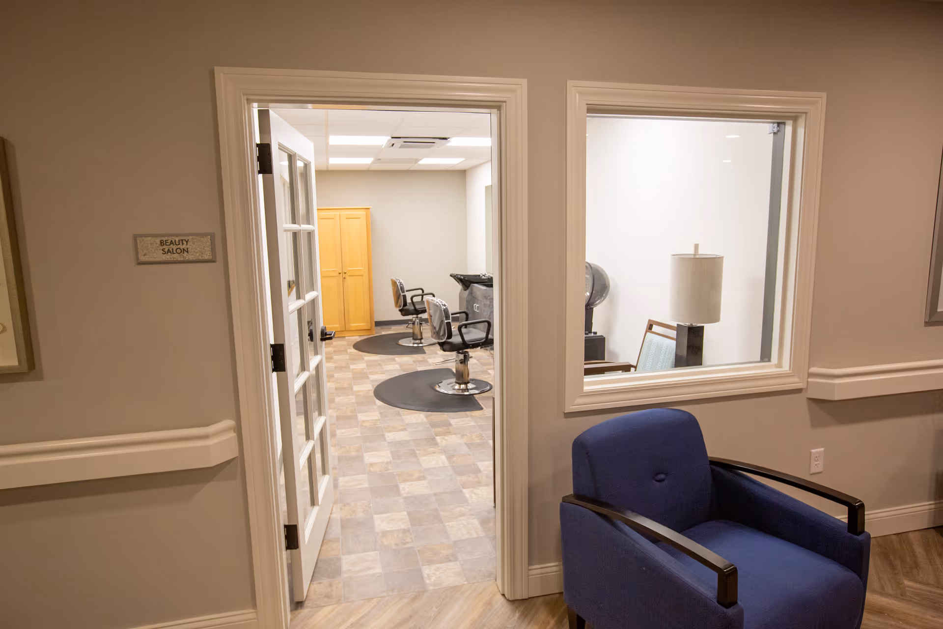 View of a beauty salon inside a senior living facility. The image shows an open door leading to the salon with two salon chairs on black mats, a wooden cabinet, and a hair washing station. In the foreground, there is a blue armchair and a window with a lamp and chair visible inside the salon. The walls are painted light gray with white trim.