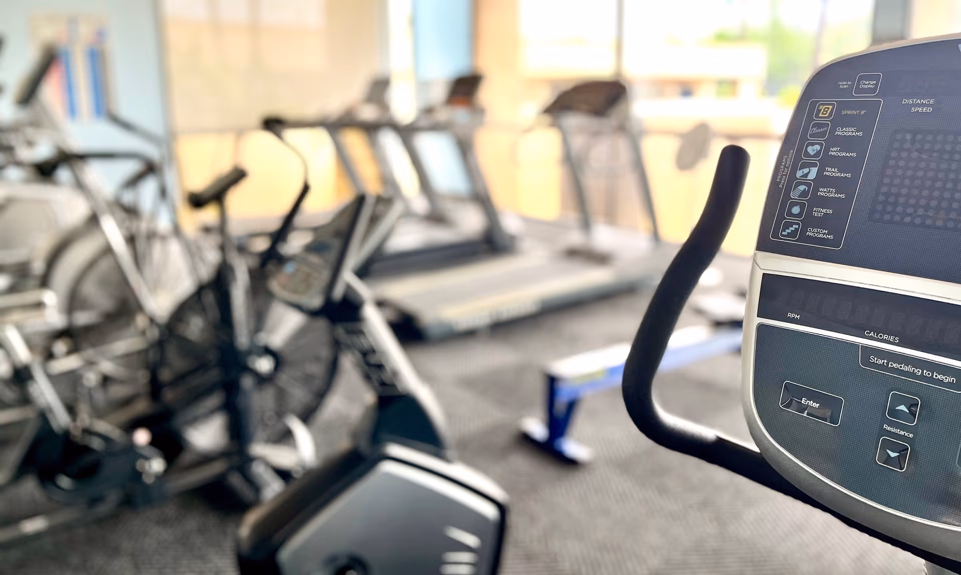 Indoor fitness room with exercise equipment including stationary bikes and treadmills, with a focus on the control panel of a stationary bike.