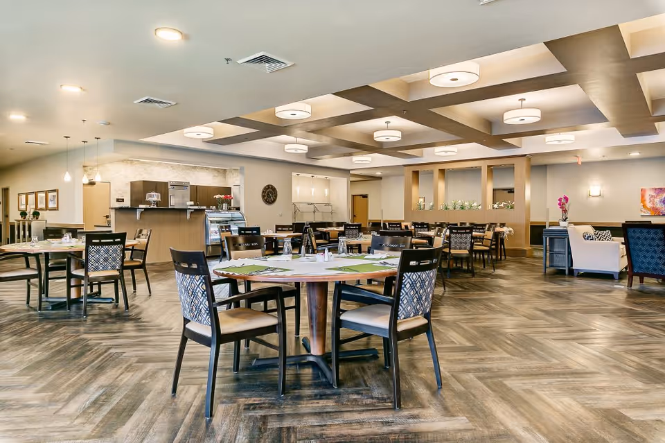 A spacious dining room in a senior living facility with multiple round and rectangular tables surrounded by chairs. The room features a modern ceiling design with recessed lighting and pendant lights over a counter area. There is a small kitchen or serving area in the background with a display case and coffee machine. The floor has a wood-patterned finish, and there are decorative elements like flowers and artwork on the walls.