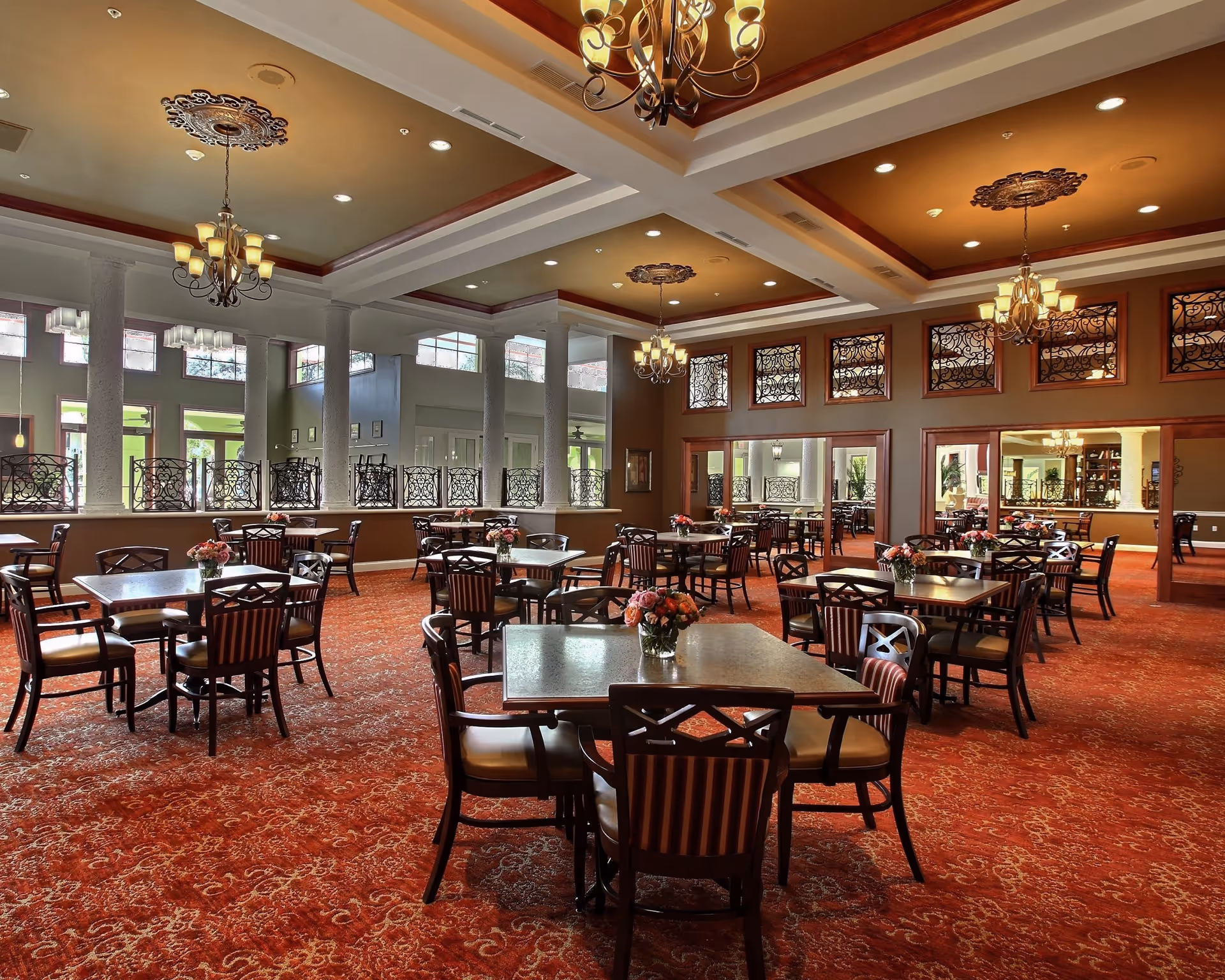 A spacious dining room with multiple square tables and chairs arranged neatly on a red patterned carpet. Each table has a small floral centerpiece. The room features large windows with decorative ironwork, white columns, and elegant chandeliers hanging from a coffered ceiling.