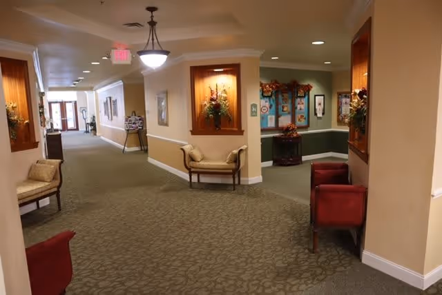 A carpeted hallway in a senior living facility with beige walls and ceiling lights. There are decorative flower arrangements in recessed wall niches and a few chairs placed along the walls. At the end of the hallway, there are glass doors letting in natural light. A bulletin board with autumn-themed decorations is visible in a side alcove.