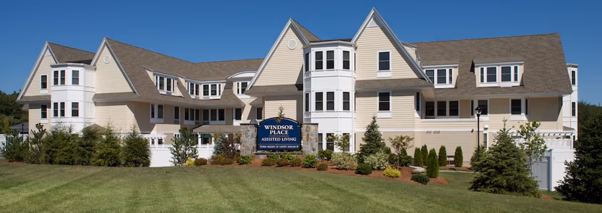Exterior view of a large, beige assisted living facility building with multiple windows and peaked roofs under a clear blue sky. The building is surrounded by landscaped bushes and a well-maintained lawn. A sign in front reads 'Windsor Place Assisted Living'.