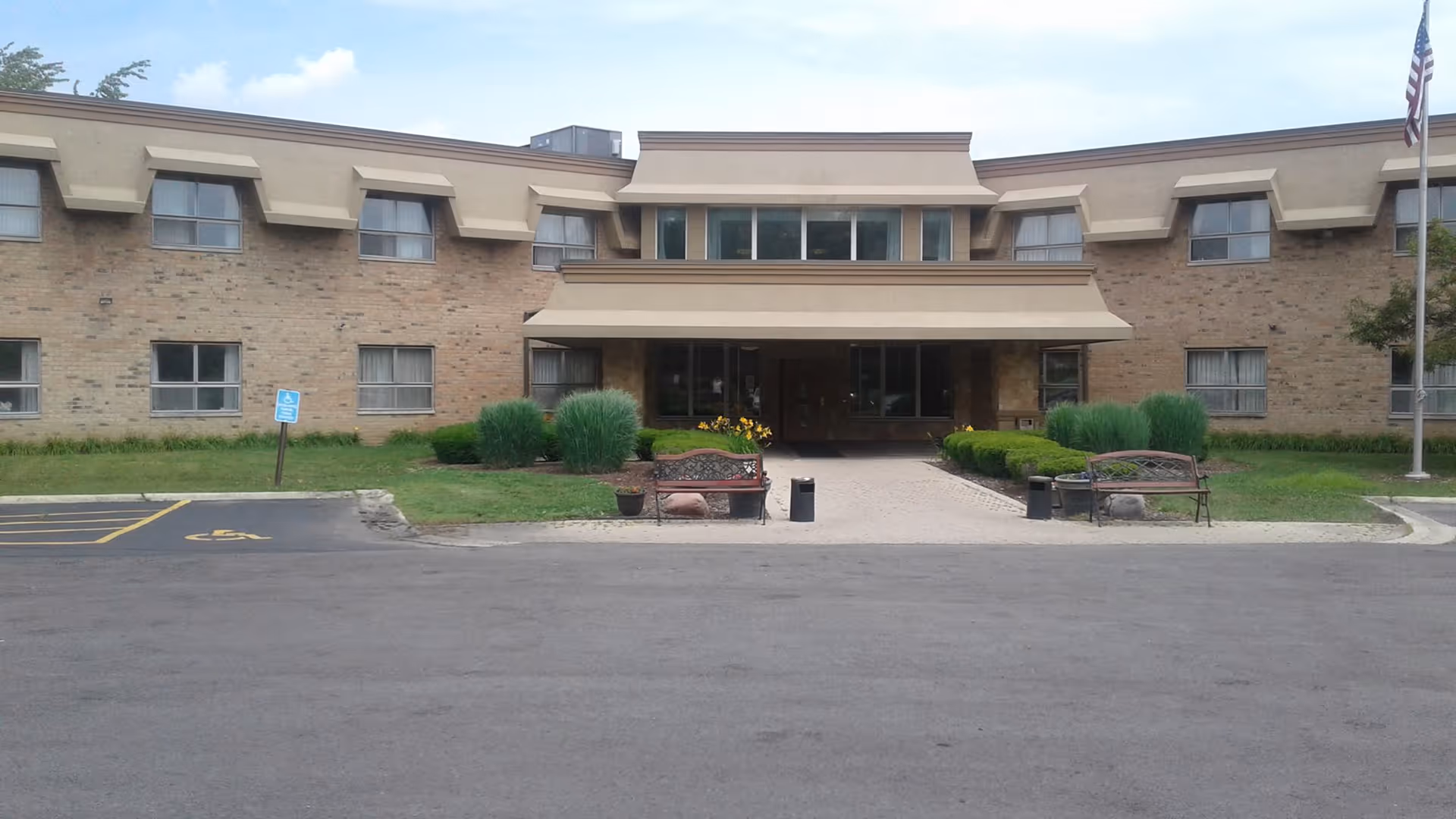 Front exterior view of a two-story brick building with multiple windows, a covered entrance with benches and landscaping, a flagpole with an American flag, and a parking area with a handicap parking space.