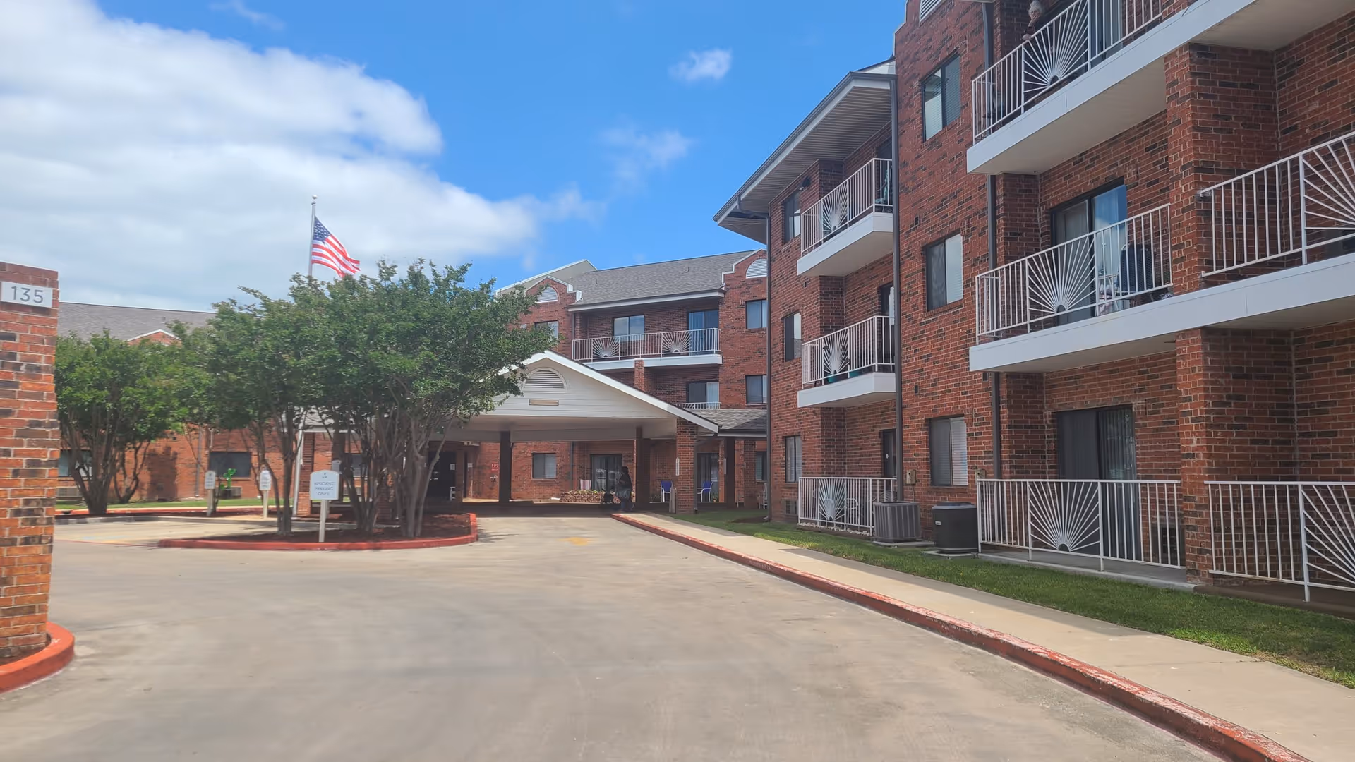 Driveway and covered entrance of a multi-story brick senior living building with balconies and an American flag.