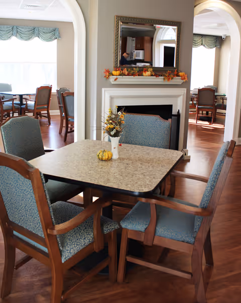 A dining area with a square table surrounded by four wooden chairs with blue patterned cushions. The table has a small vase with flowers and a decorative pumpkin. In the background, there is a white fireplace with a mirror above it, decorated with autumn leaves. The room has wooden flooring and arched doorways leading to more tables and chairs near large windows with green valances.