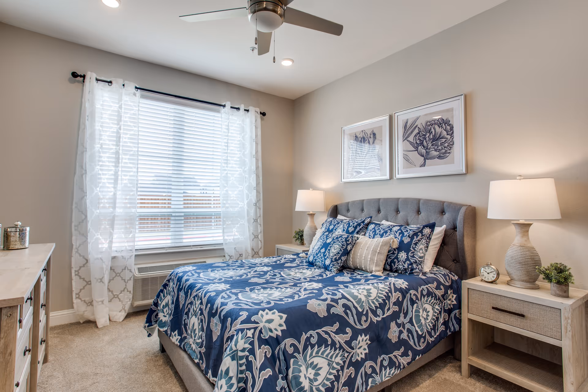 A cozy bedroom with a large window covered by white patterned curtains and blinds. The bed has a tufted gray headboard and is dressed in blue and white floral bedding with multiple pillows. There are two matching bedside tables, each with a lamp, a small plant, and an alarm clock on one side. The walls are painted light beige and decorated with two framed floral prints above the bed. A ceiling fan with lights is mounted on the ceiling.