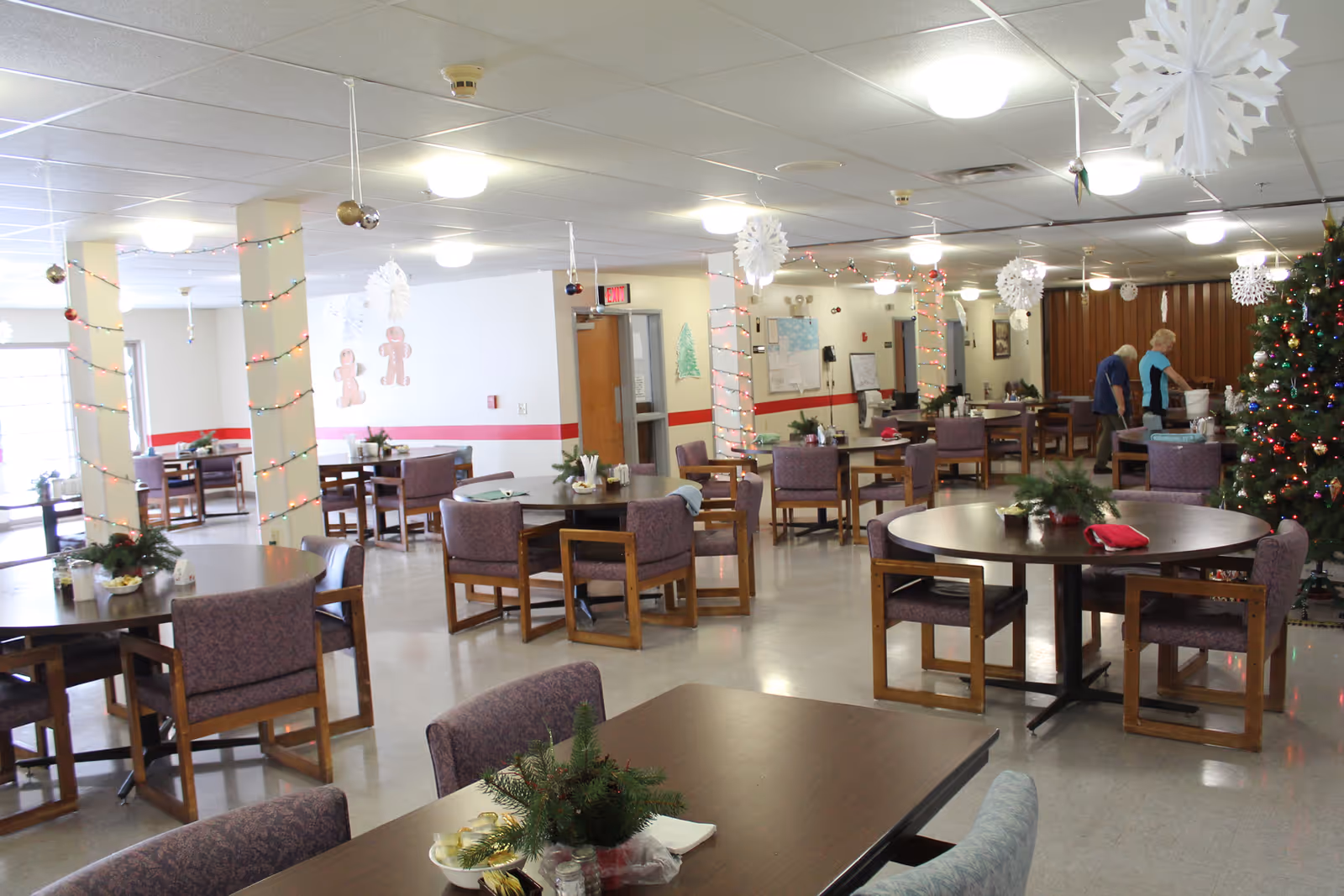 A spacious dining room decorated for the holidays with Christmas lights wrapped around pillars, hanging paper snowflakes, and a decorated Christmas tree. Several round and rectangular tables with chairs are arranged throughout the room. Two elderly women are seen near the back of the room, one standing and the other bending over a bucket. The walls have holiday decorations including gingerbread men and a green Christmas tree cutout.