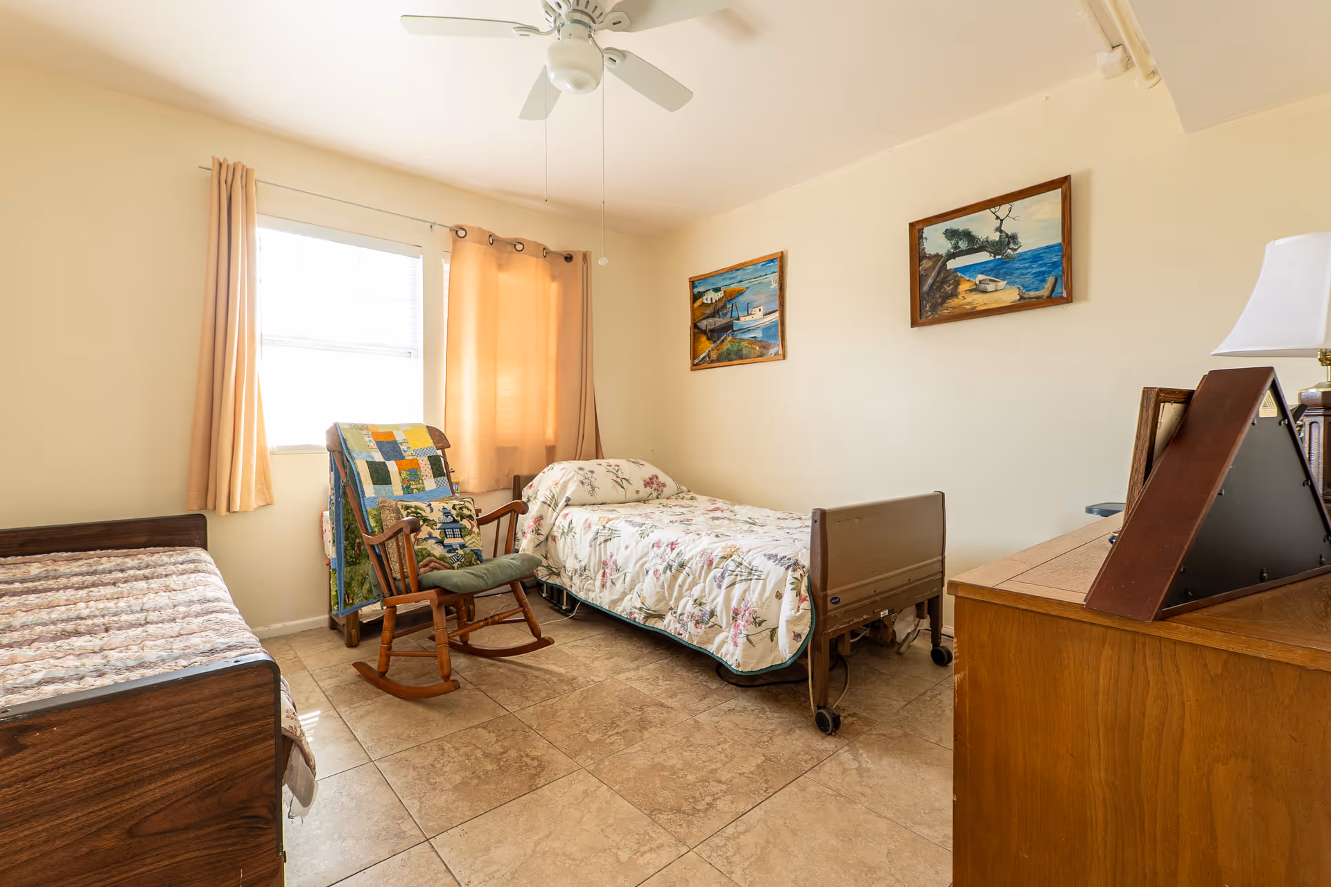A senior living bedroom with two beds, one with a floral bedspread and the other with a patterned quilt. There is a wooden rocking chair with a cushion and a colorful quilt draped over it near a window with beige curtains. The room has tiled floors, two paintings on the wall, a ceiling fan, and a wooden dresser with a lamp and a photo frame.