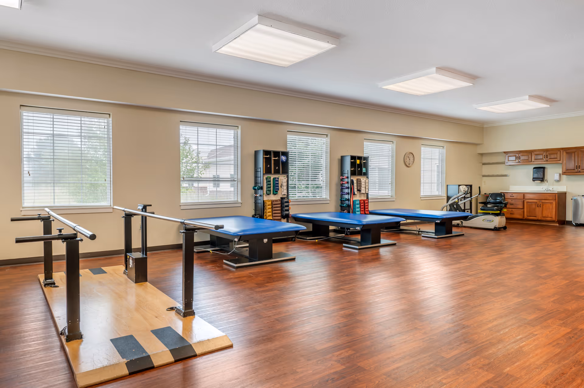 A spacious physical therapy room with wooden flooring, three blue therapy tables, parallel bars for walking exercises, racks with various exercise bands and weights, a stationary exercise bike, and a sink with wooden cabinets. The room has several windows with blinds allowing natural light to enter.