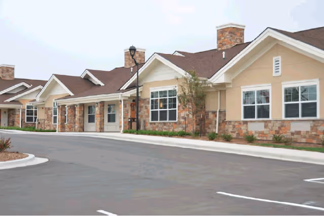 Exterior view of a single-story senior living facility building with beige walls, stone accents, multiple windows, and a brown roof under a cloudy sky. The building is adjacent to a paved parking lot with marked parking spaces.