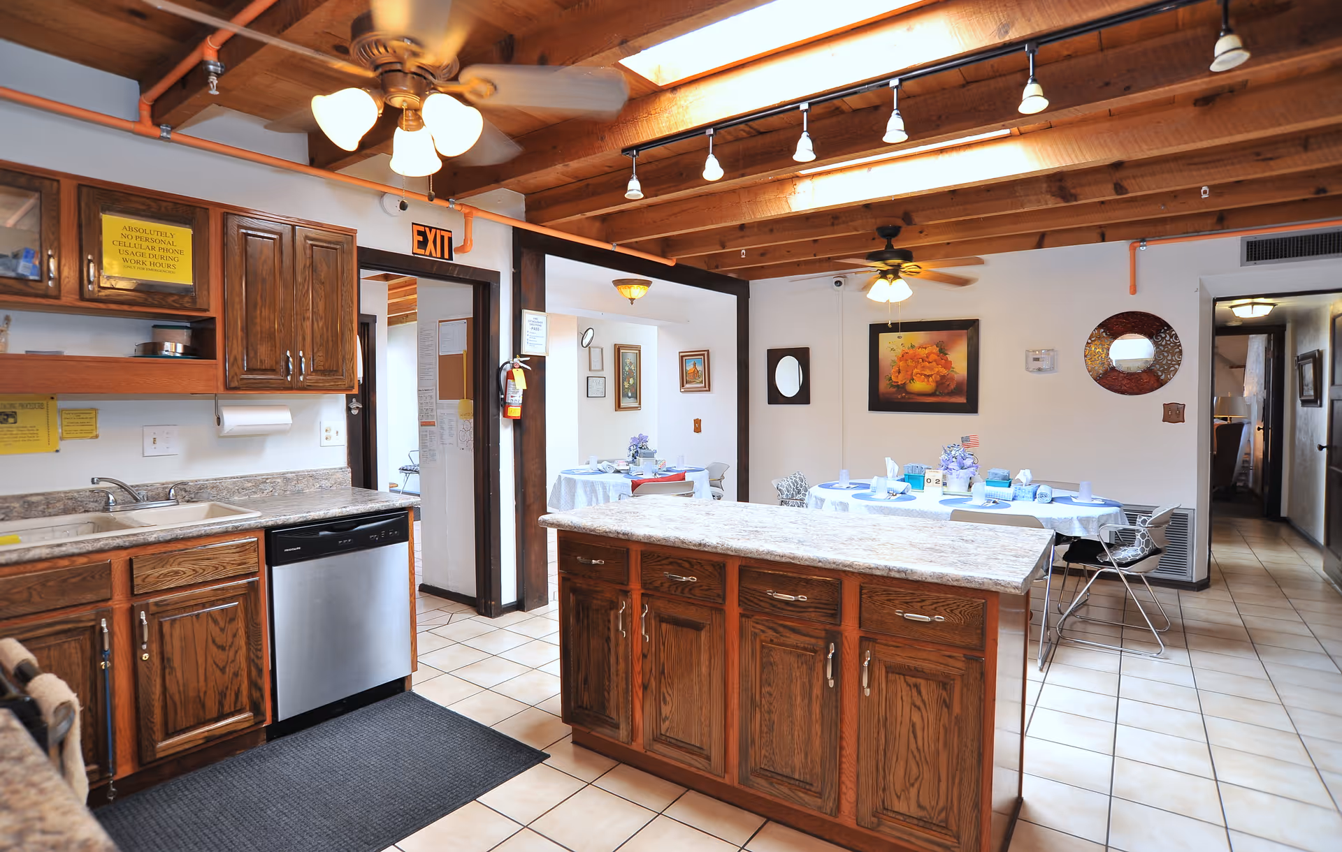 Interior view of a kitchen and dining area in an assisted living facility. The kitchen features wooden cabinets, a dishwasher, a sink, and a central island with a granite countertop. The dining area has round tables covered with white tablecloths, set with plates, cups, and napkins. The ceiling has exposed wooden beams with ceiling fans and track lighting. There are framed pictures and a decorative mirror on the walls.