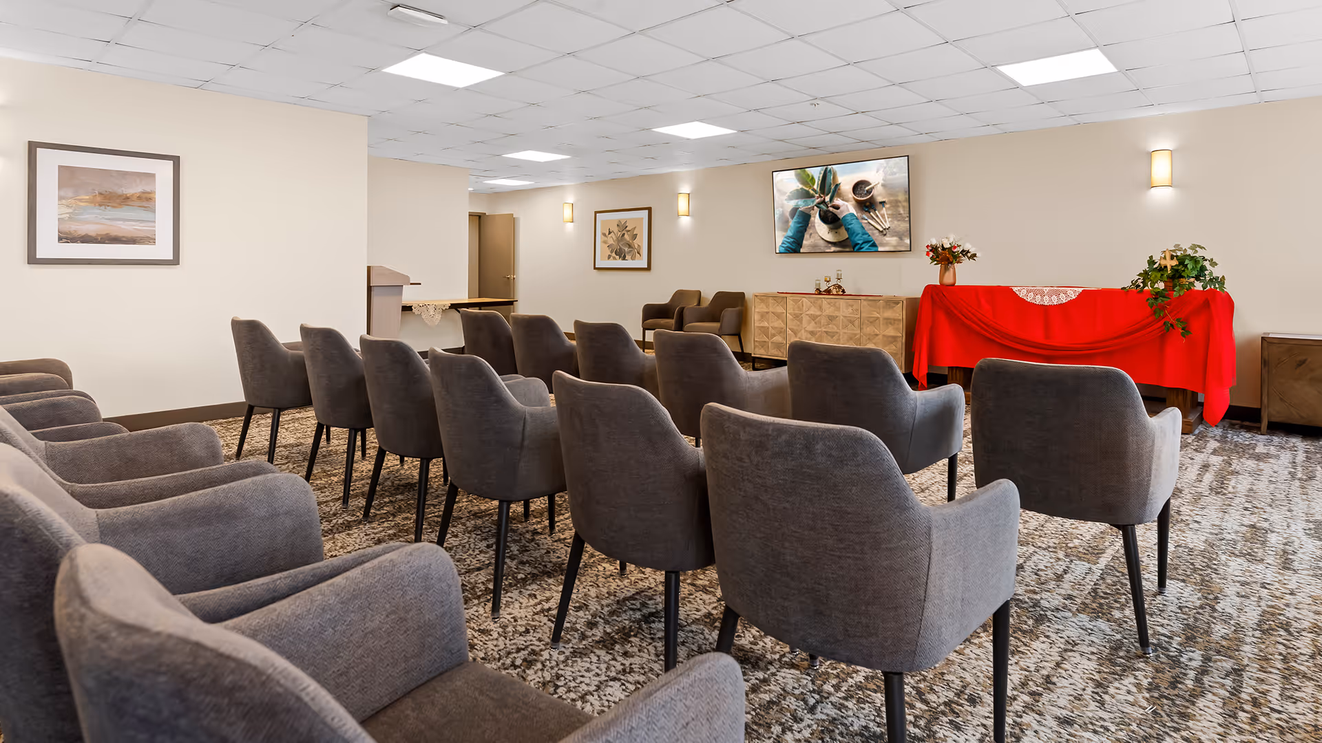 A meeting or presentation room with multiple rows of gray upholstered chairs facing a wall-mounted TV screen. The room has beige walls, carpeted floor, framed artwork on the walls, a wooden podium, and a table covered with a red cloth and decorated with plants and flowers.