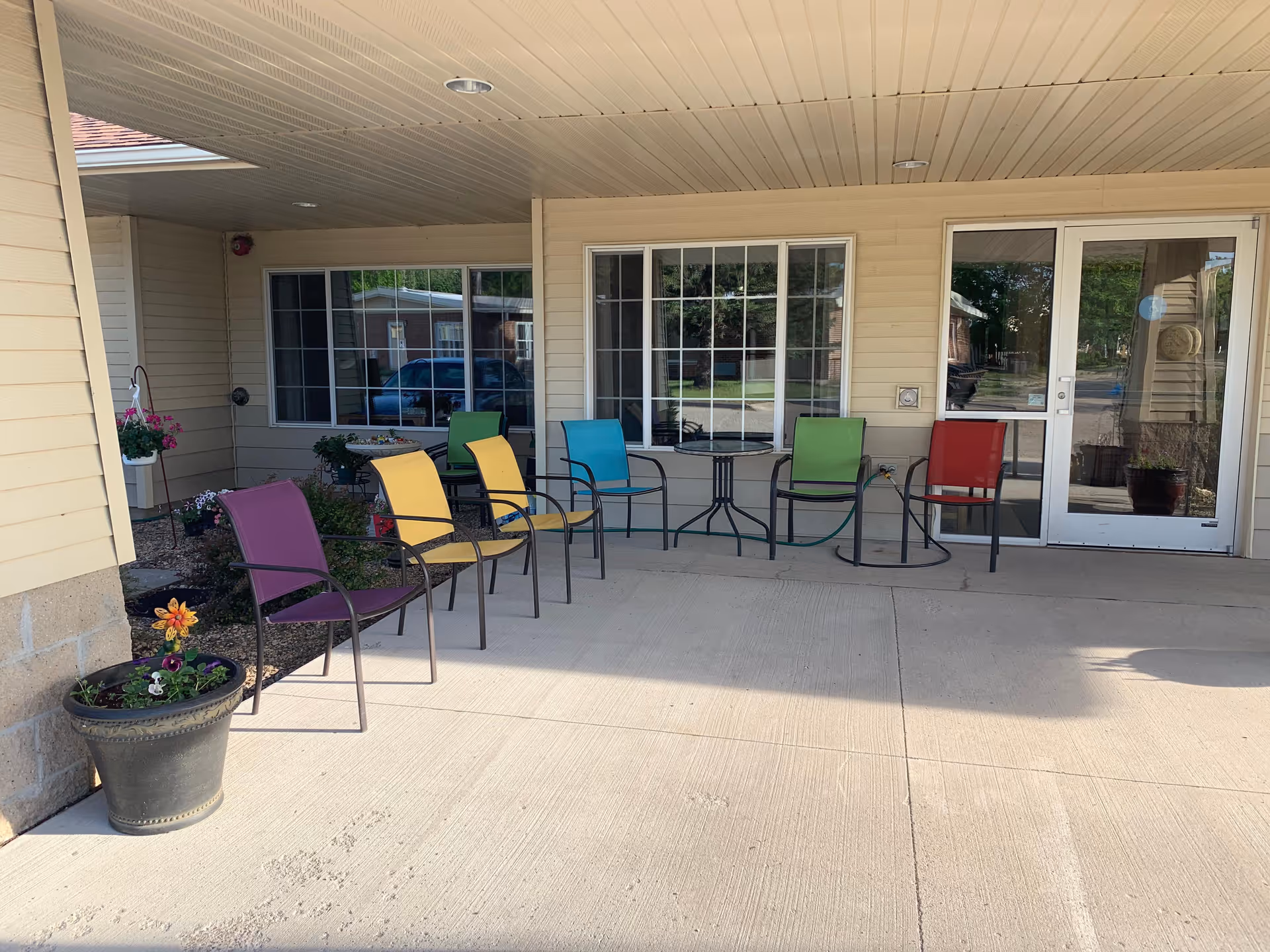 Covered entrance patio with a row of colorful outdoor chairs, potted plants, windows, and a glass door.