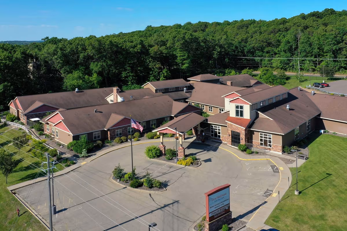 Aerial view of Dove Healthcare – South Assisted Living facility surrounded by greenery. The building has a brown roof and beige walls with red accents. There is a circular driveway with an American flag in the center and a sign near the entrance. Parking spaces and a wooded area are visible in the background.