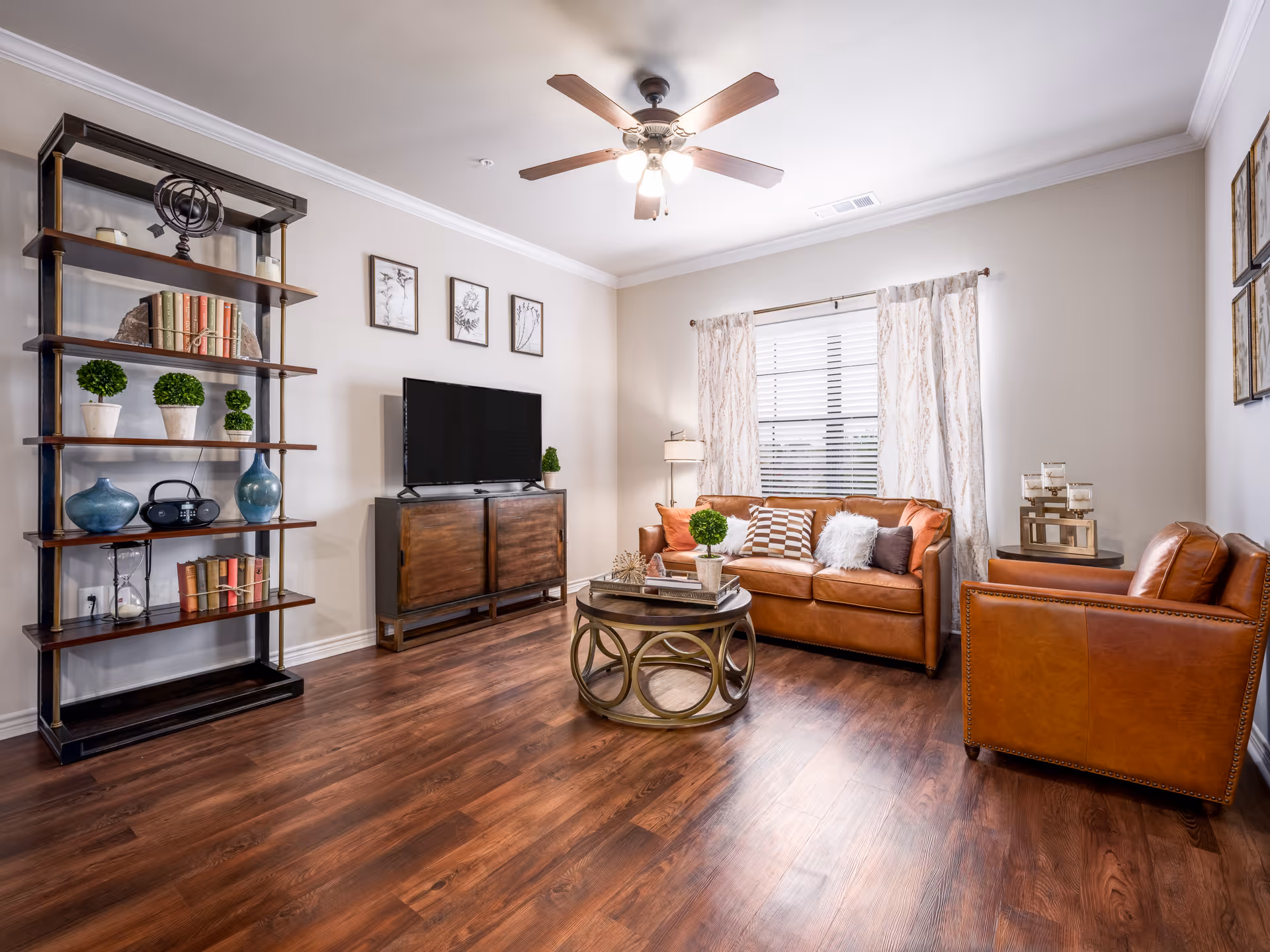 A cozy living room with wooden flooring, a brown leather sofa and matching armchair, a round coffee table with decorative items, a wooden TV stand with a flat-screen TV, a tall bookshelf with books and decorative plants, and a ceiling fan with lights. The room has light-colored walls and a window with patterned curtains.