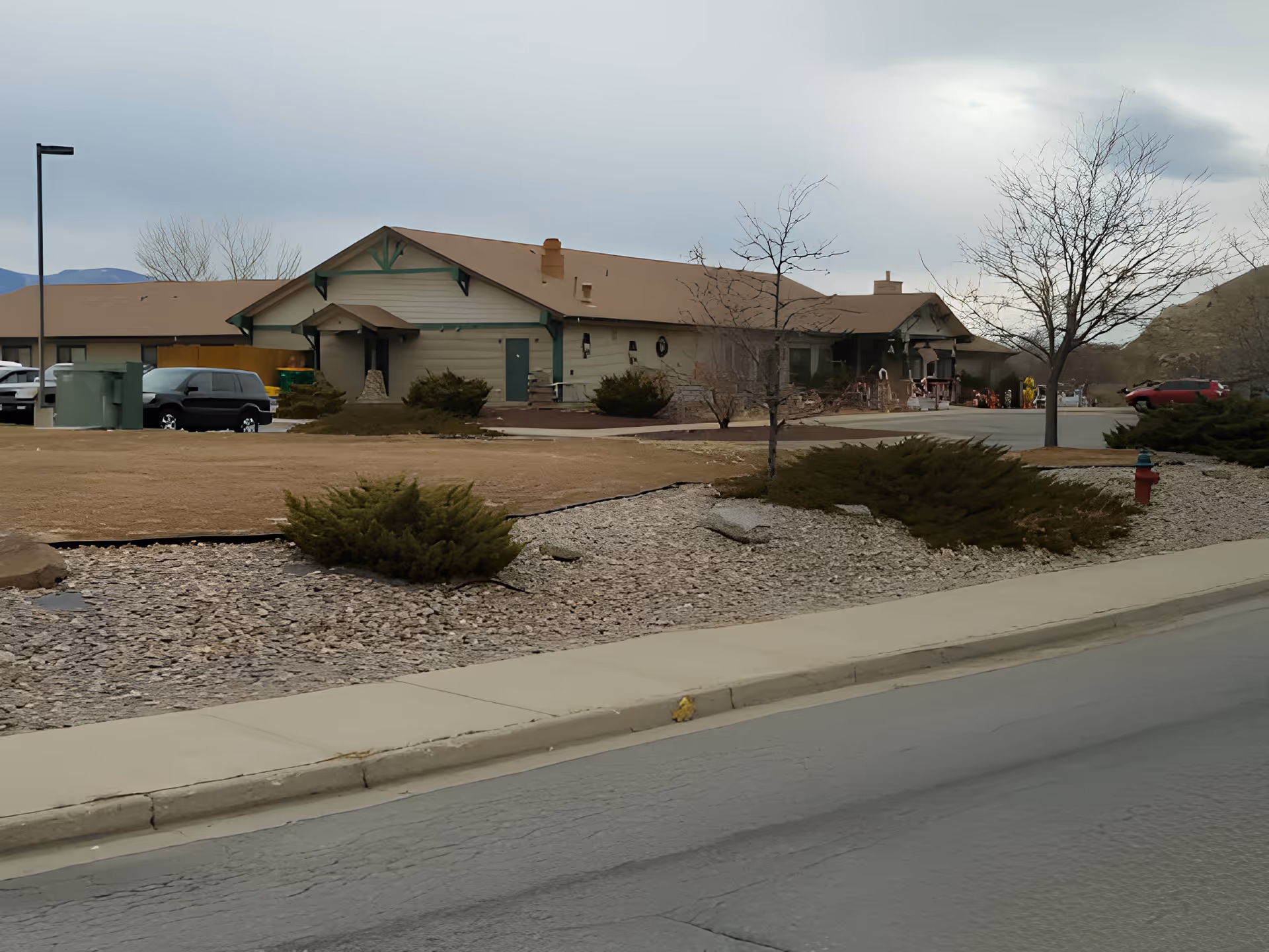 Exterior view of a single-story building with a brown roof and beige walls, surrounded by a landscaped area with bushes, leafless trees, and a rock garden. Several parked cars are visible near the building, and a fire hydrant is seen on the right side near the road. The sky is overcast.