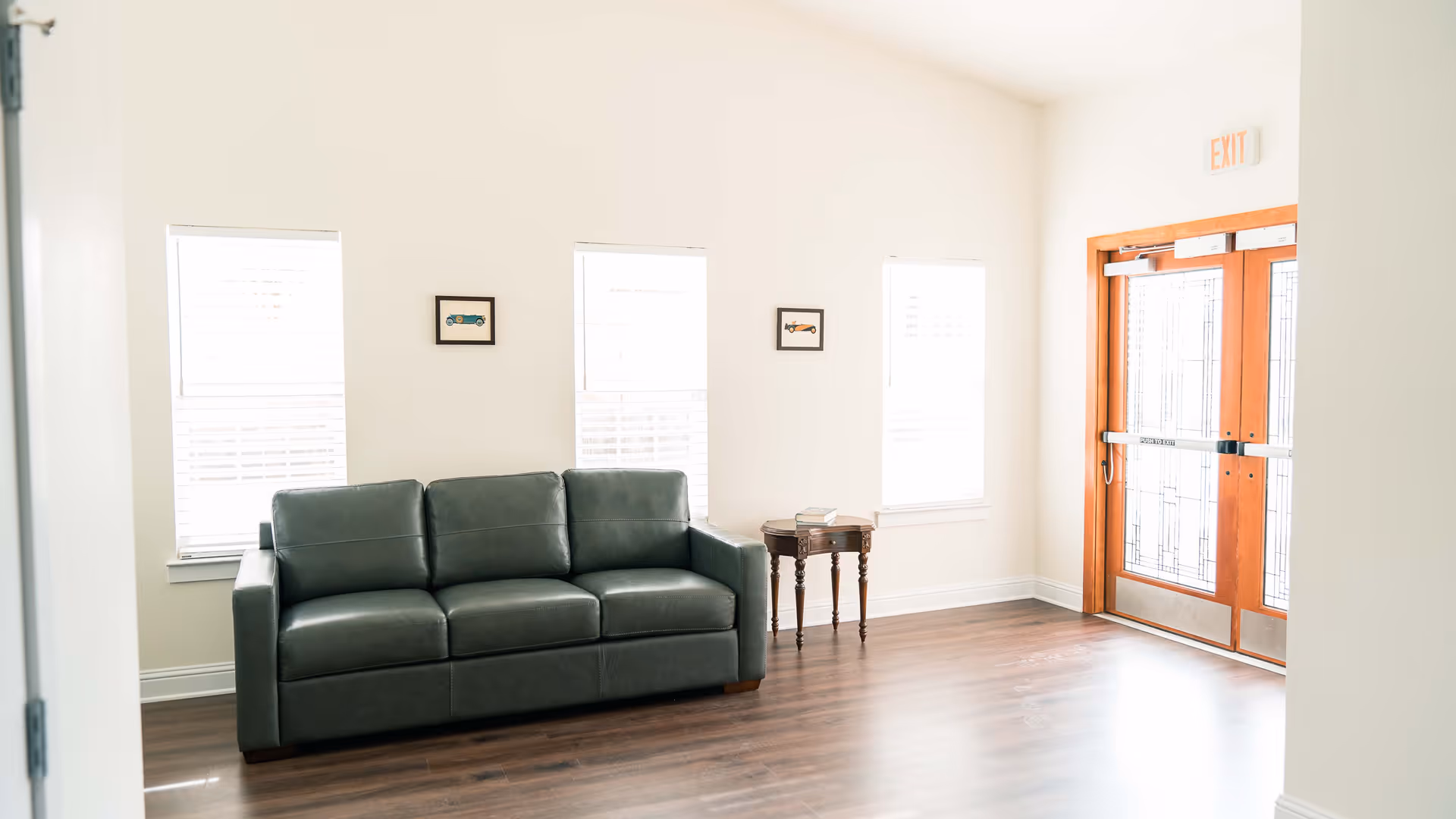 A bright room with three windows and a wooden floor featuring a dark green leather sofa and a small wooden side table with books on it. There are two small framed pictures on the wall above the sofa, and a double wooden door with glass panels and an exit sign above it.