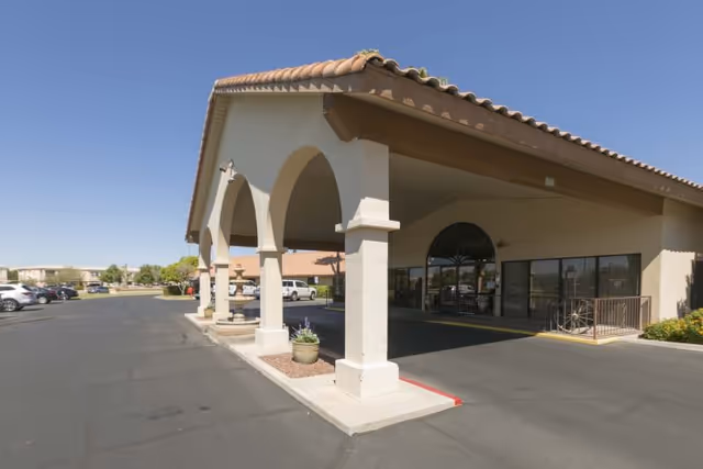 Exterior view of the entrance to Lake Pleasant Post Acute Rehabilitation Center featuring a covered drop-off area with arches and a tiled roof, adjacent parking lot, and clear blue sky.