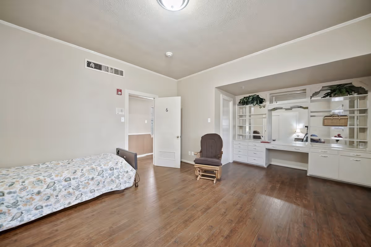 A simple bedroom with a single bed covered in a floral bedspread, a wooden rocking chair, and a large built-in white dresser with mirrors and shelves. The room has wooden flooring, beige walls, and a door marked with the number 6 that is slightly open.