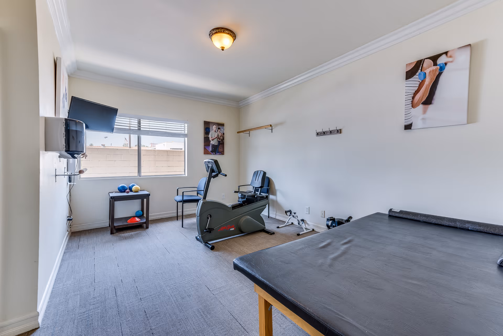 A small exercise room with a stationary recumbent bike, two chairs, a small table with exercise balls, and a padded exercise table. The room has a window with blinds, a wall-mounted TV, and two fitness-themed pictures on the walls.