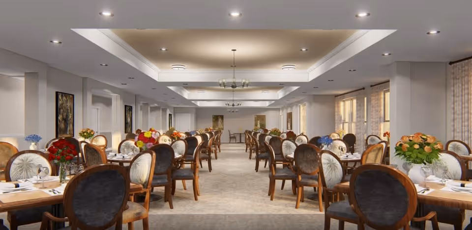 A spacious dining room with multiple wooden tables and cushioned chairs arranged in rows. Each table is decorated with colorful floral centerpieces and set with plates, glasses, and silverware. The room has a neutral color palette with soft lighting from ceiling fixtures and large windows with curtains on the right side.