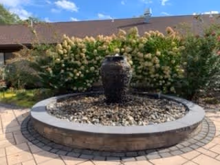 Outdoor circular stone water fountain surrounded by a paved patio area with green bushes and a building with a brown roof in the background under a blue sky with some clouds.