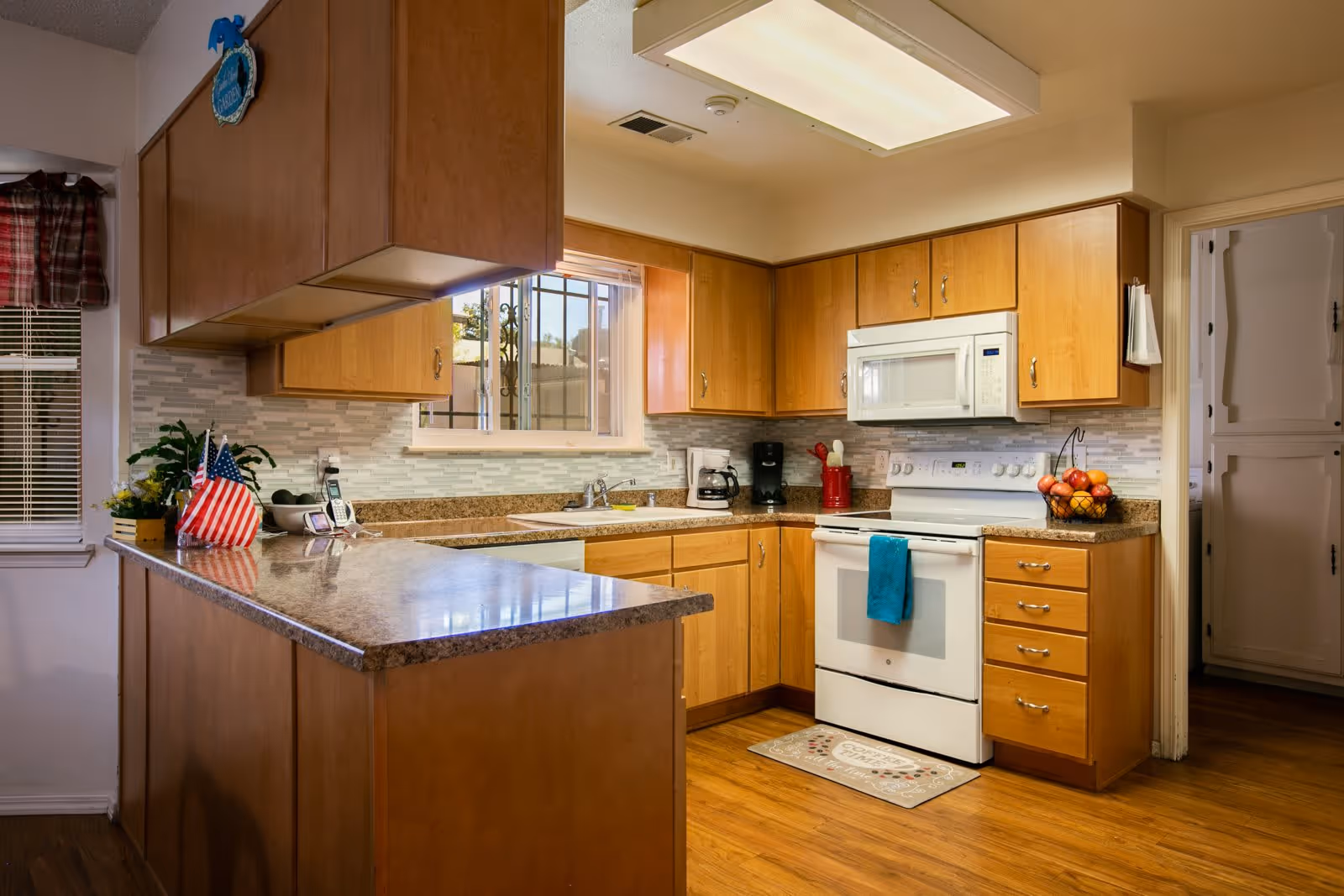 A well-lit kitchen with wooden cabinets, a white stove with a blue towel hanging on the handle, a white microwave above the stove, a coffee maker, a basket of fruit on the counter, and a window above the sink. The kitchen has a granite countertop and wooden flooring.