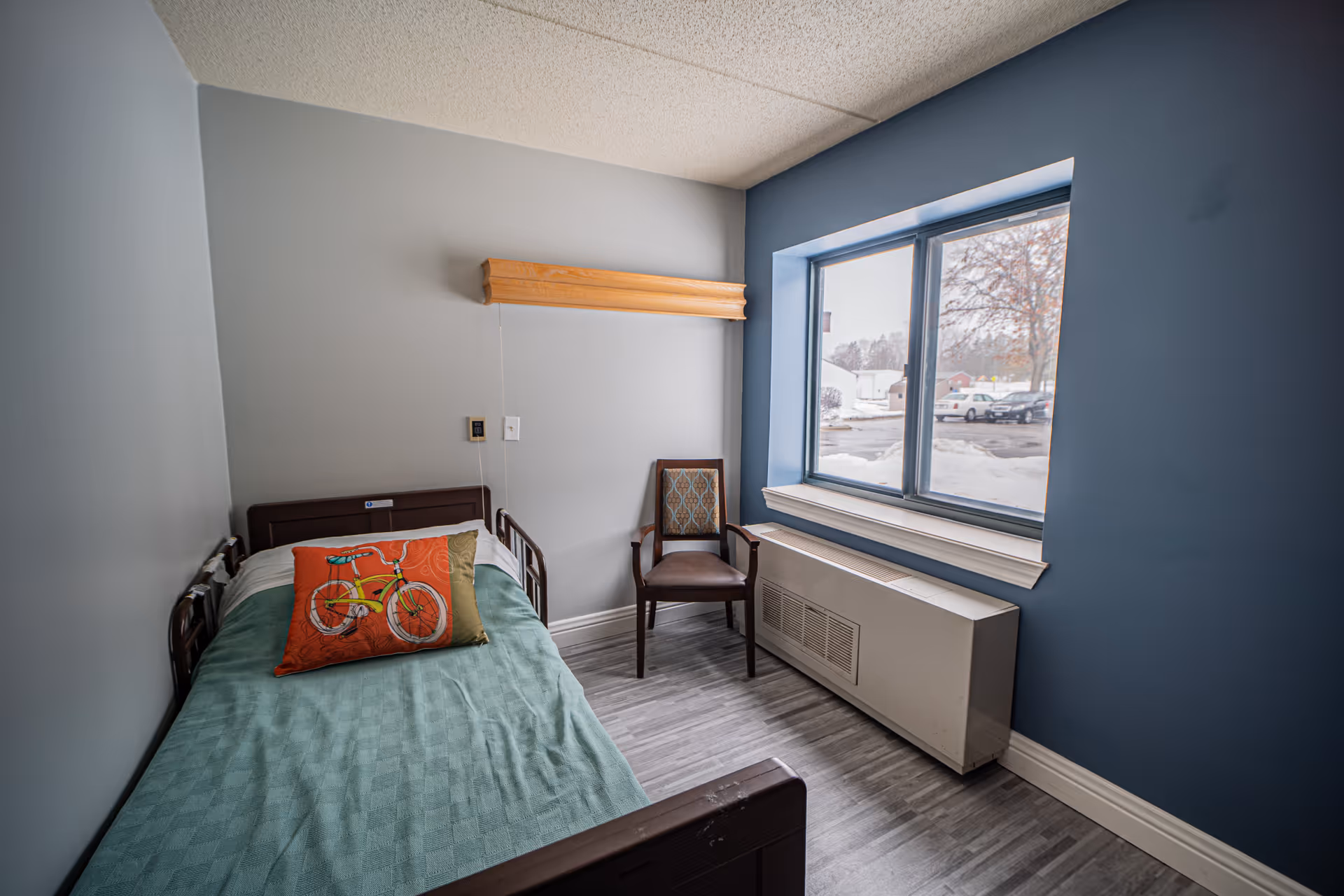 A small bedroom with a single bed covered with a teal blanket and two pillows, one with a bicycle design. There is a wooden chair with patterned upholstery next to a large window showing a snowy outdoor scene. The walls are painted gray and blue, and the floor has gray wood-like flooring.