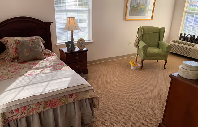 A cozy bedroom with a bed covered in floral bedding, a wooden nightstand with a lamp and picture frames, a green upholstered armchair near a window, and a dresser with decorative boxes. The room has beige carpet and white walls with a framed picture hanging.