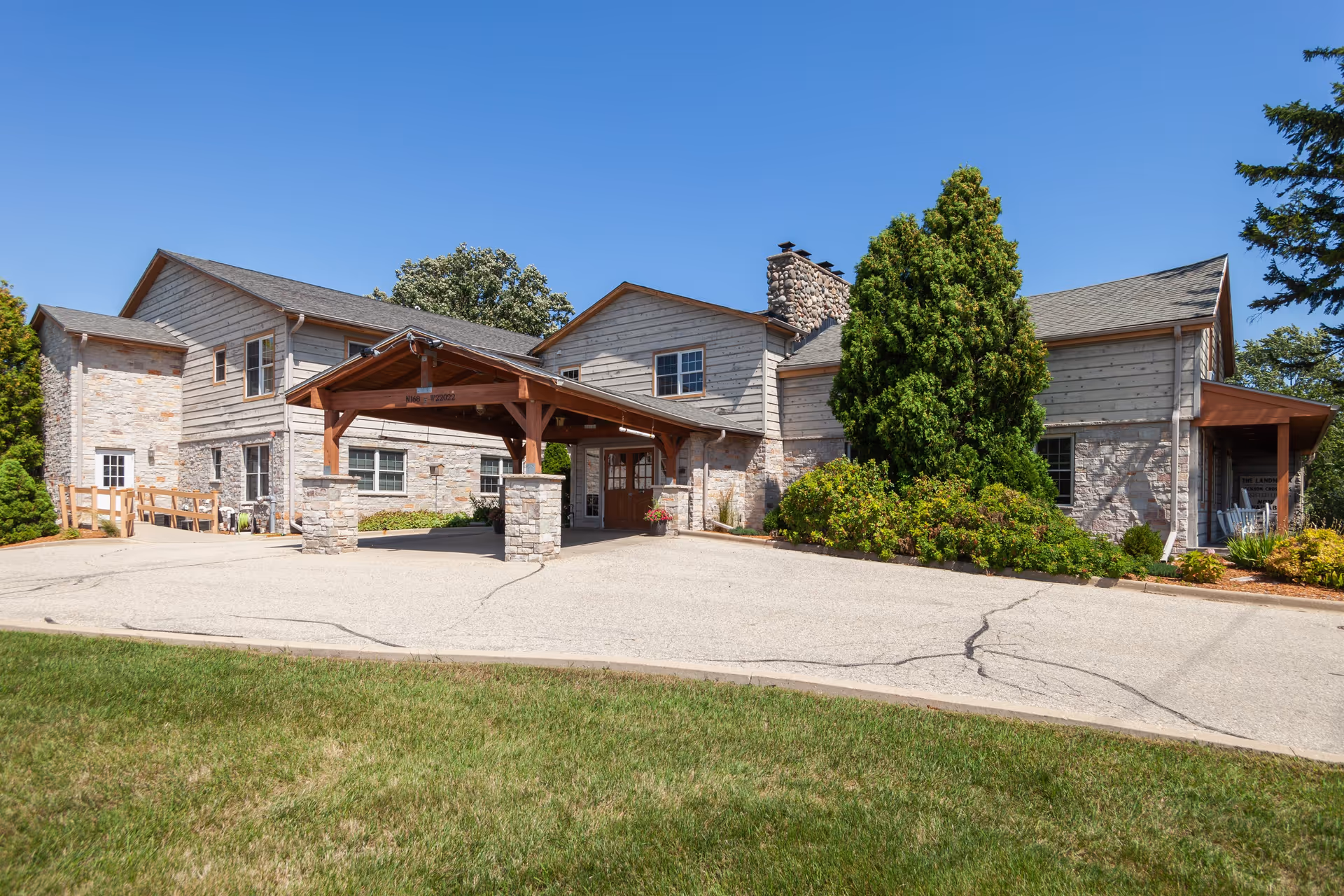 Front exterior of a two-story senior living building with a covered drop-off entrance, stone and wood siding, and surrounding landscaping.