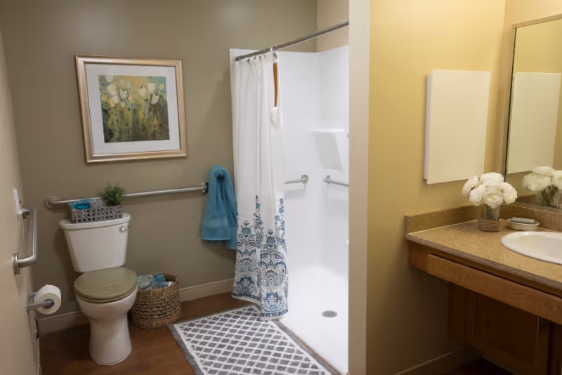 A clean and well-lit bathroom featuring a toilet with a green seat cover, a wicker basket with towels, a wall-mounted grab bar, a shower with a white curtain decorated with blue patterns, a gray and white patterned rug, a countertop with a sink, a mirror, and a vase with white flowers. A framed floral painting hangs on the wall above the toilet.