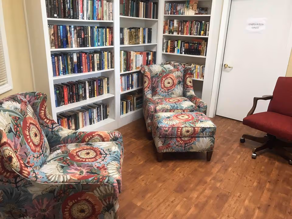 Cozy reading room with white bookshelves, two floral armchairs and an ottoman on a wood floor.