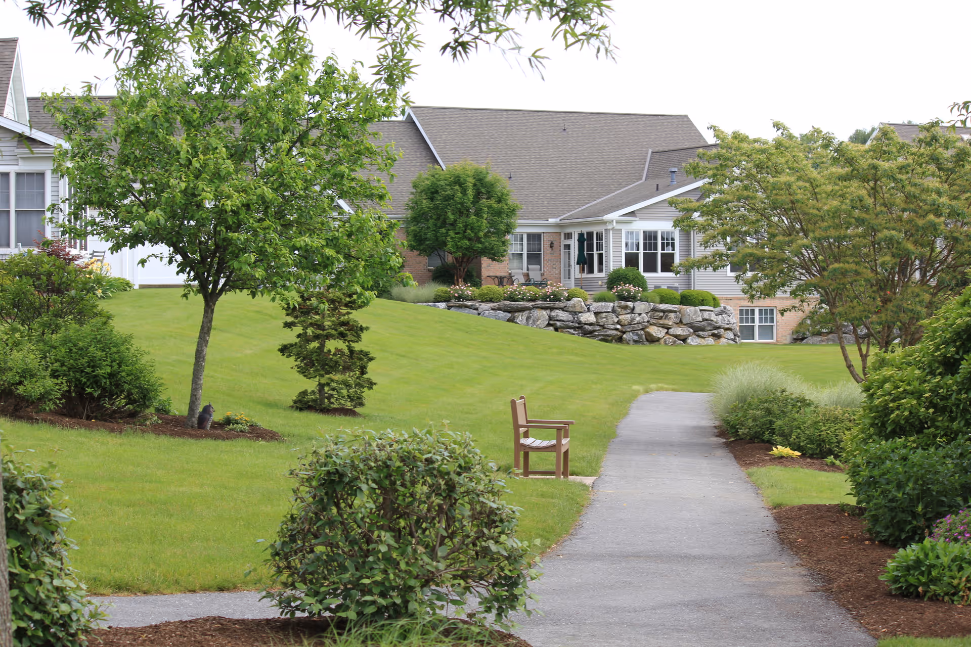 A paved walkway through landscaped lawns and shrubs leading to a bench with residential buildings in the background.