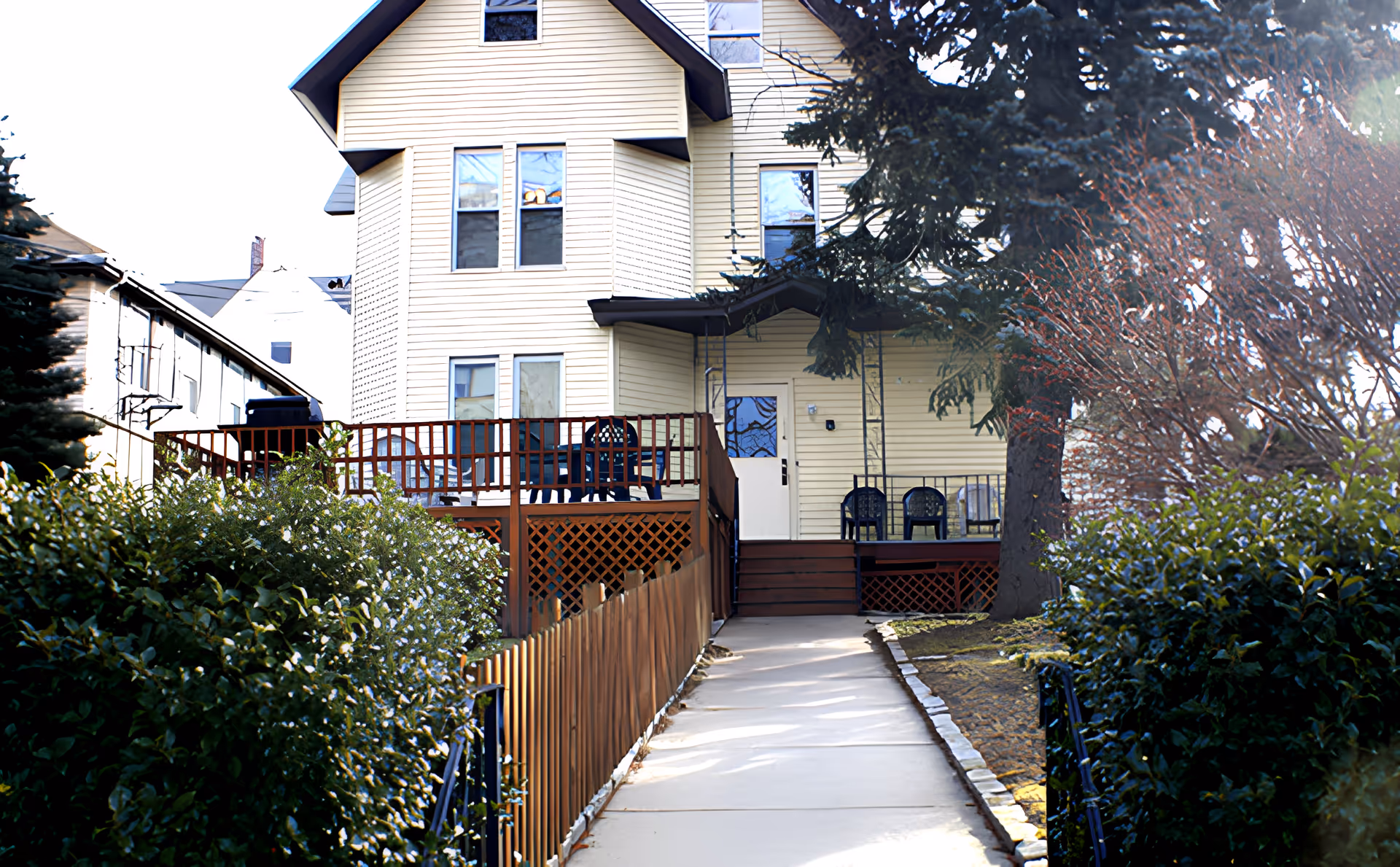 A paved walkway leads to the entrance of a multi-story beige building with several windows. The building has a wooden deck with outdoor chairs and a railing. There are bushes and trees on either side of the walkway.