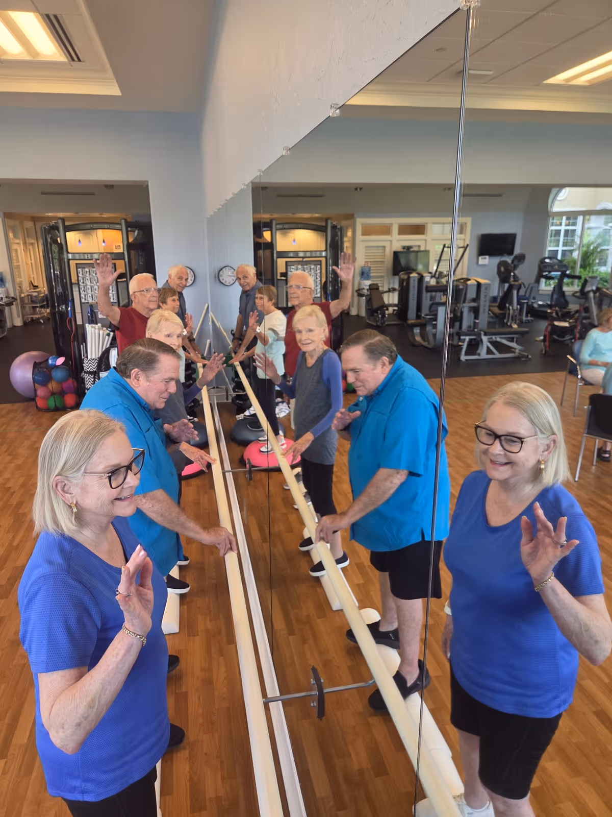 A group of elderly people participating in a fitness or exercise class in a gym room. They are standing along a ballet barre, facing a large mirror, and waving or raising their hands. The room has wooden floors, exercise equipment, and large windows letting in natural light.