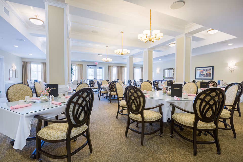 Bright assisted living dining room with round tables set for meals, upholstered chairs, and chandeliers.