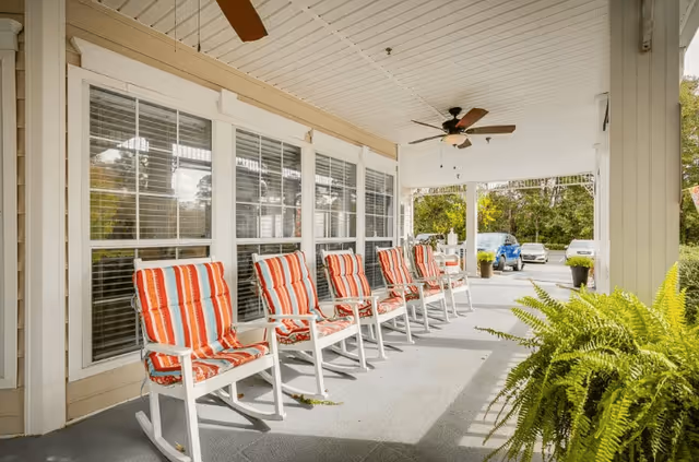 Covered outdoor porch area with six white rocking chairs featuring red and white striped cushions lined up against a wall with large windows. Ceiling fans are mounted on the white ceiling, and there are green plants and parked cars visible in the background.