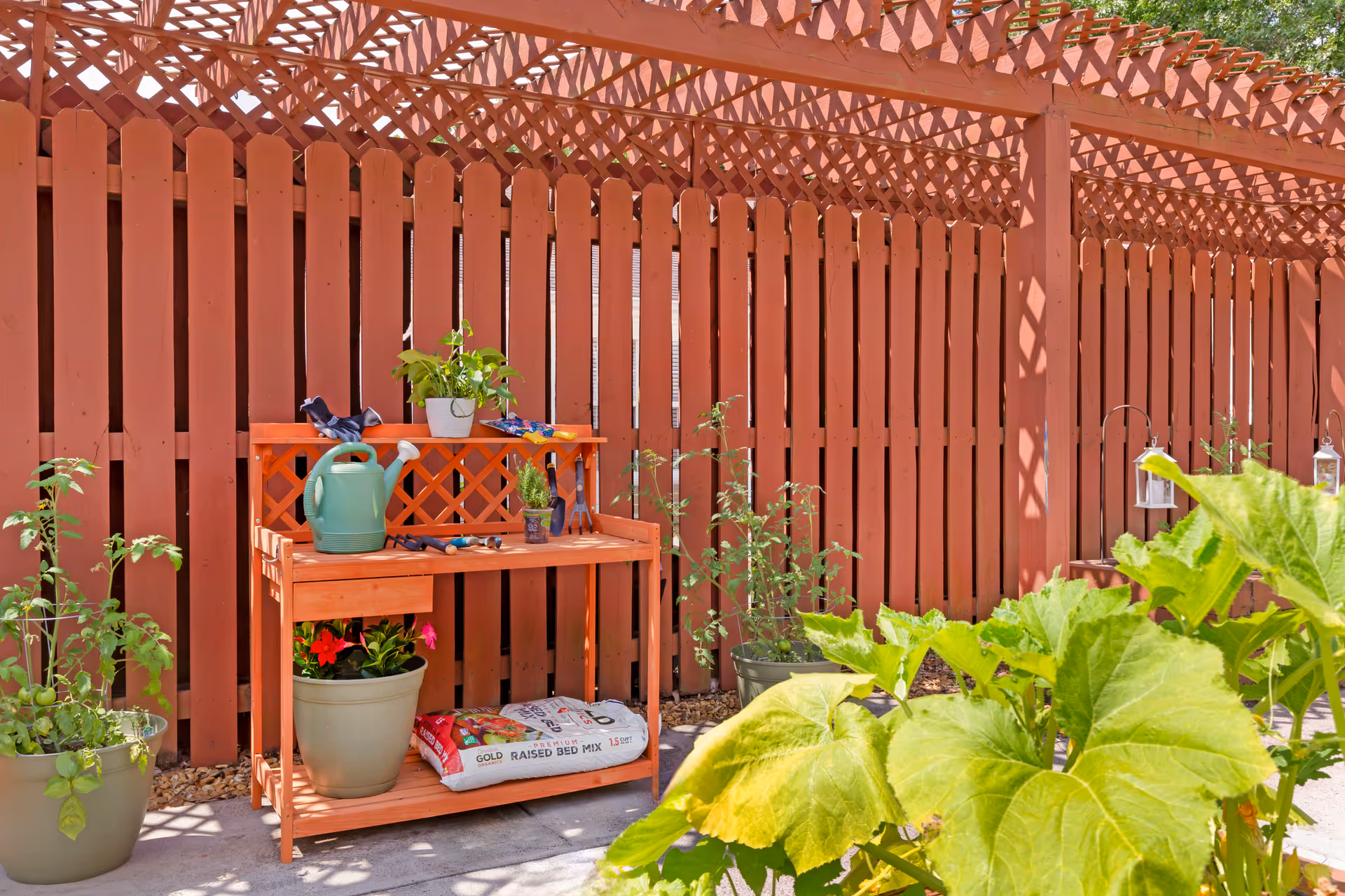 Outdoor gardening area with a wooden potting bench, watering can, potted plants, and a lattice fence under a pergola.