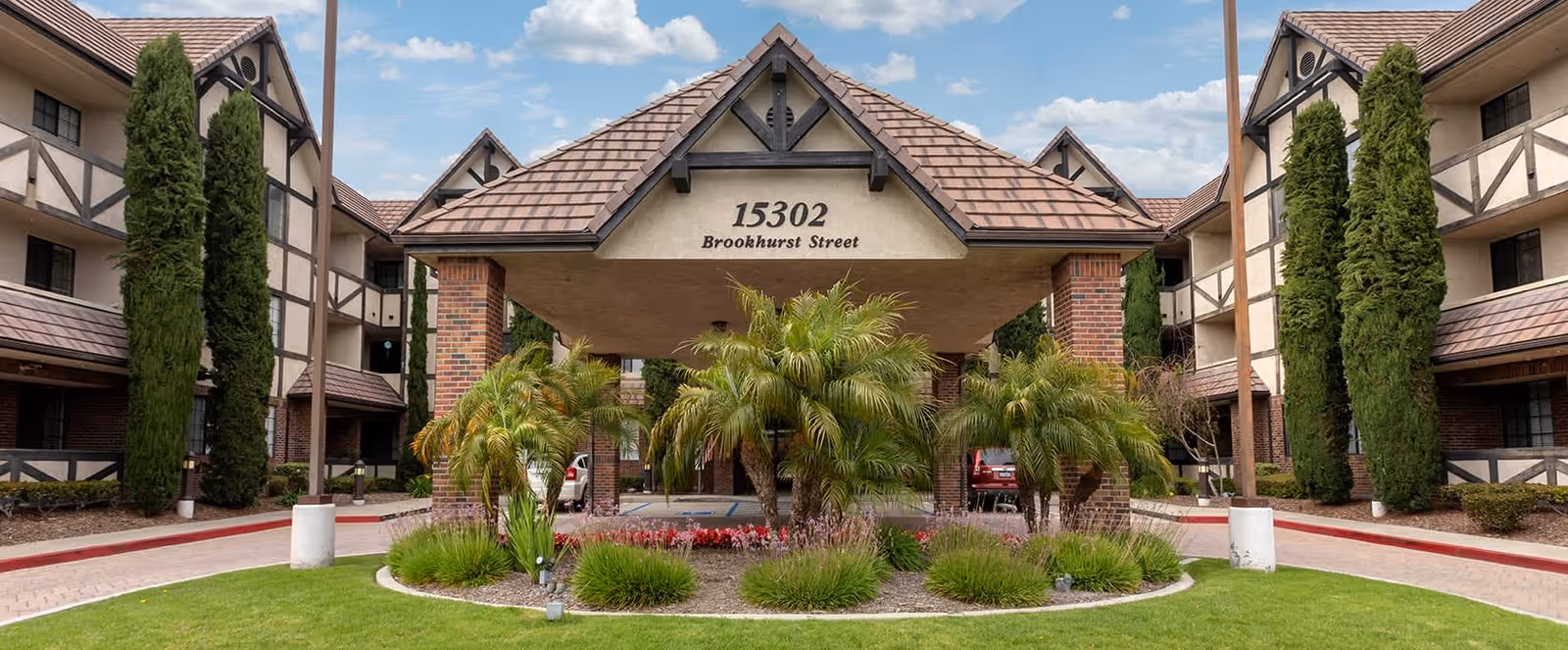 Front exterior view of Brookdale Brookhurst facility showing a covered entrance with the address 15302 Brookhurst Street, surrounded by landscaped greenery including palm trees and tall cypress trees, with a clear blue sky above.