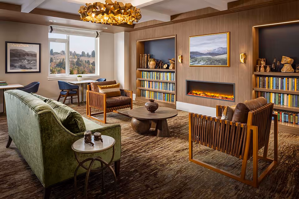 A cozy senior living room with a green velvet sofa, two wooden armchairs with leather cushions, a round wooden coffee table, built-in bookshelves filled with books and decorative items, a modern electric fireplace, and a large window showing an outdoor landscape. The room features warm lighting and a stylish chandelier.