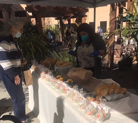 People wearing masks stand around an outdoor table with packaged snacks and pastries under a shaded pergola.