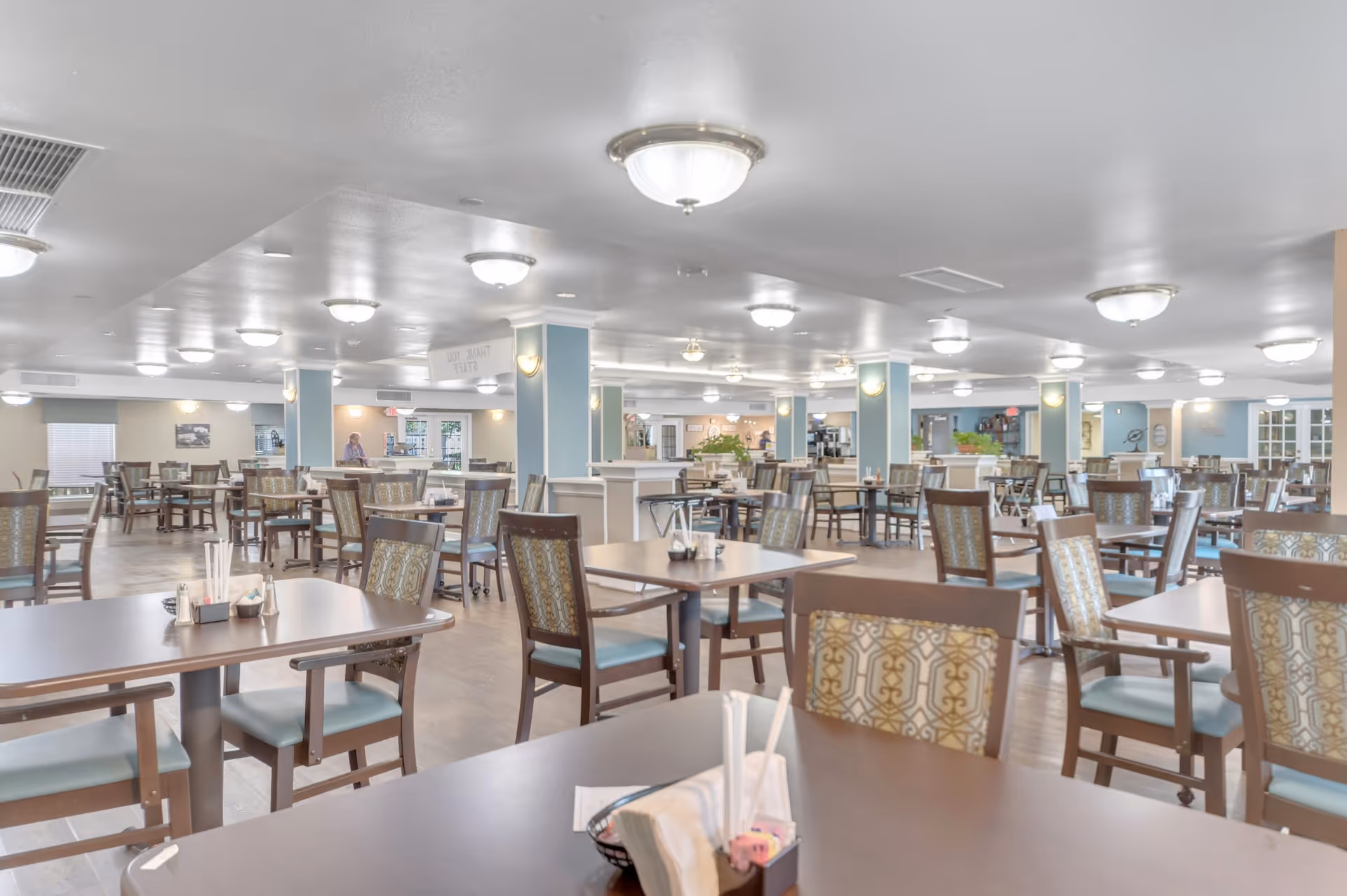 A spacious dining room with multiple wooden tables and chairs arranged neatly. The chairs have patterned upholstery and light blue cushions. The room is well-lit with ceiling lights and has light blue pillars and walls. There are some plants and a few people visible in the background near the serving area.