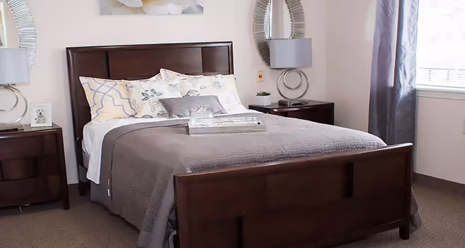 A neatly made bed with a dark wooden headboard and footboard in a bedroom. The bed has multiple pillows with floral and geometric patterns and a gray quilted bedspread. There are two matching dark wooden nightstands on either side of the bed, each with a modern lamp and decorative items. A window with gray curtains is visible on the right side of the room.